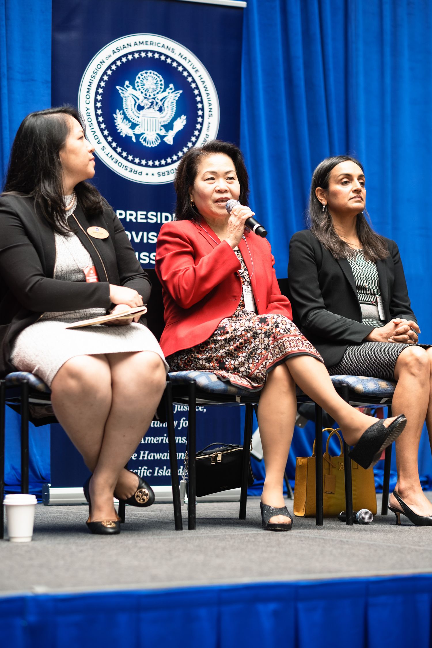Three women sit on stage at a Presidential Advisory Commission panel discussion event with blue backdrop.