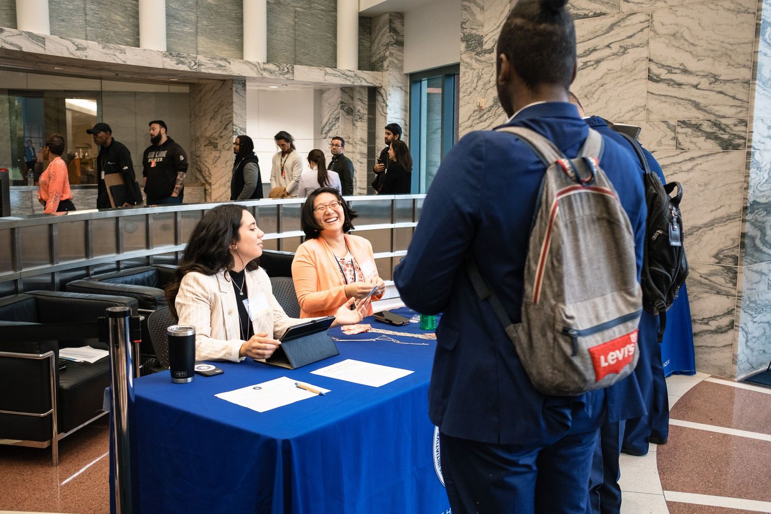 Two women at a blue registration table greeting a student with a backpack.