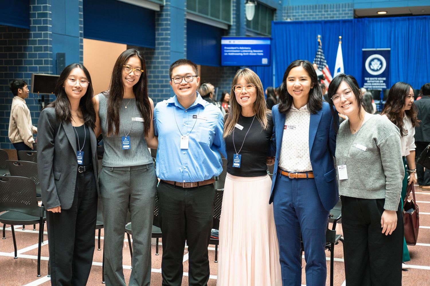 Six young Asian professionals smiling together at a formal event with blue walls and official banners in the background.