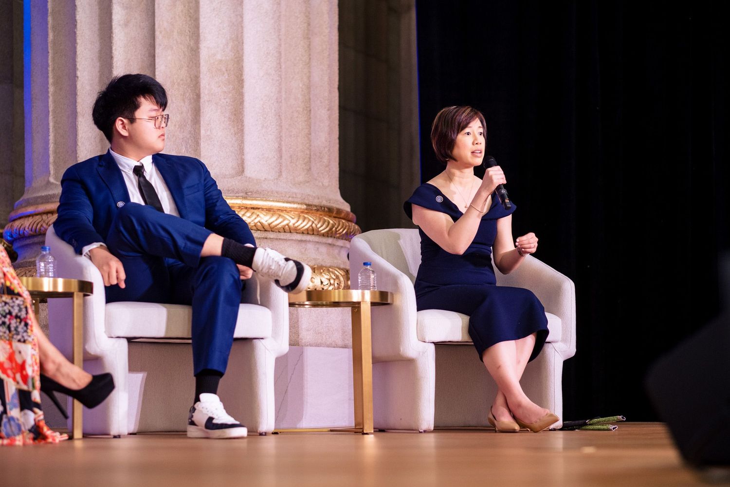Two professionals in formal attire sit on stage during a panel discussion, with a woman speaking into a microphone.