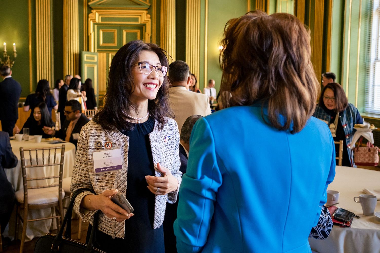 Two women networking at a formal event in an elegant green and gold ballroom, one wearing blue jacket.