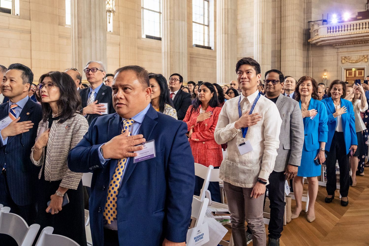 Diverse group of people reciting the Pledge of Allegiance at a formal naturalization ceremony in a grand hall.