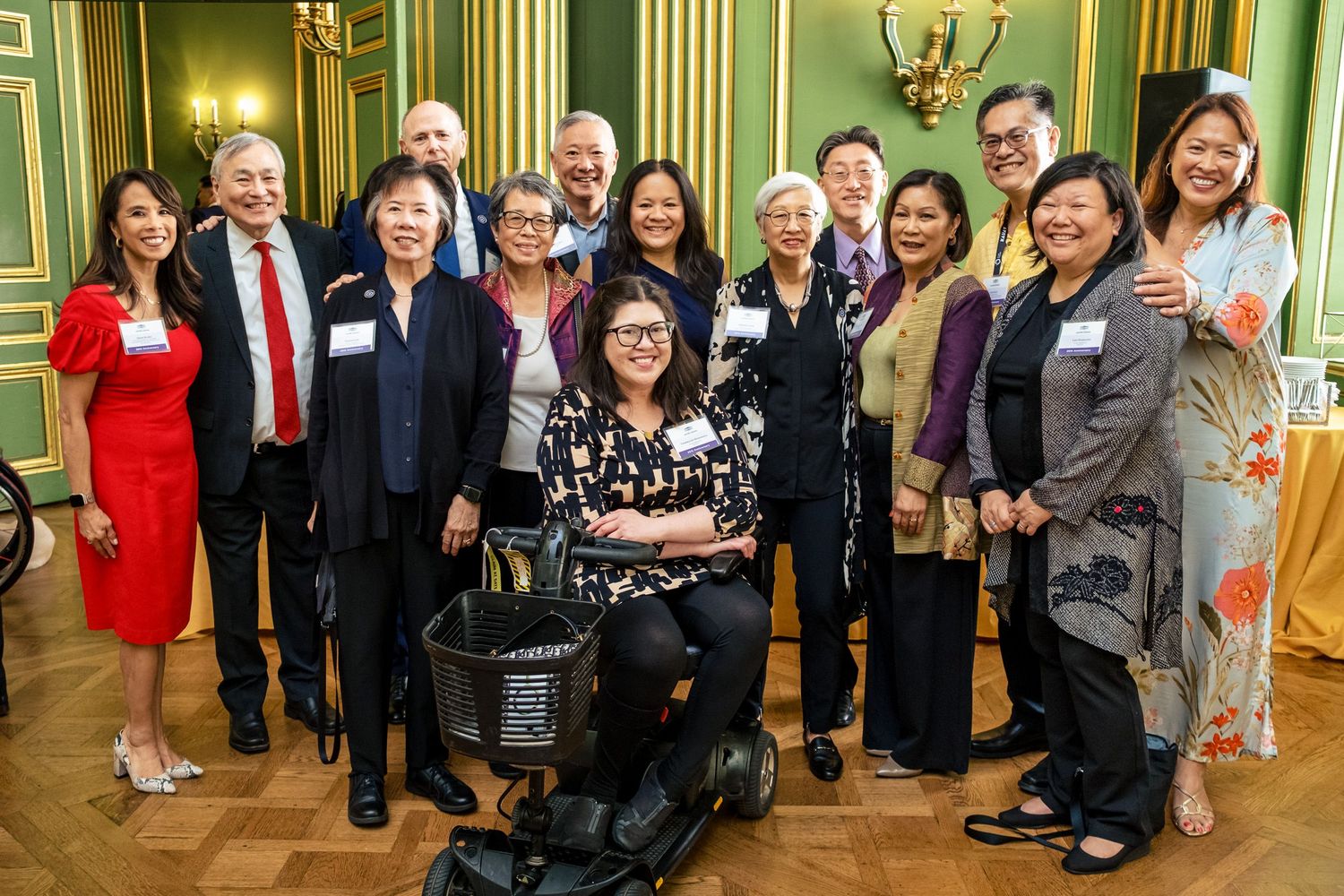Diverse group of professionals posing together in an elegant green-walled event hall, including a woman in a wheelchair.