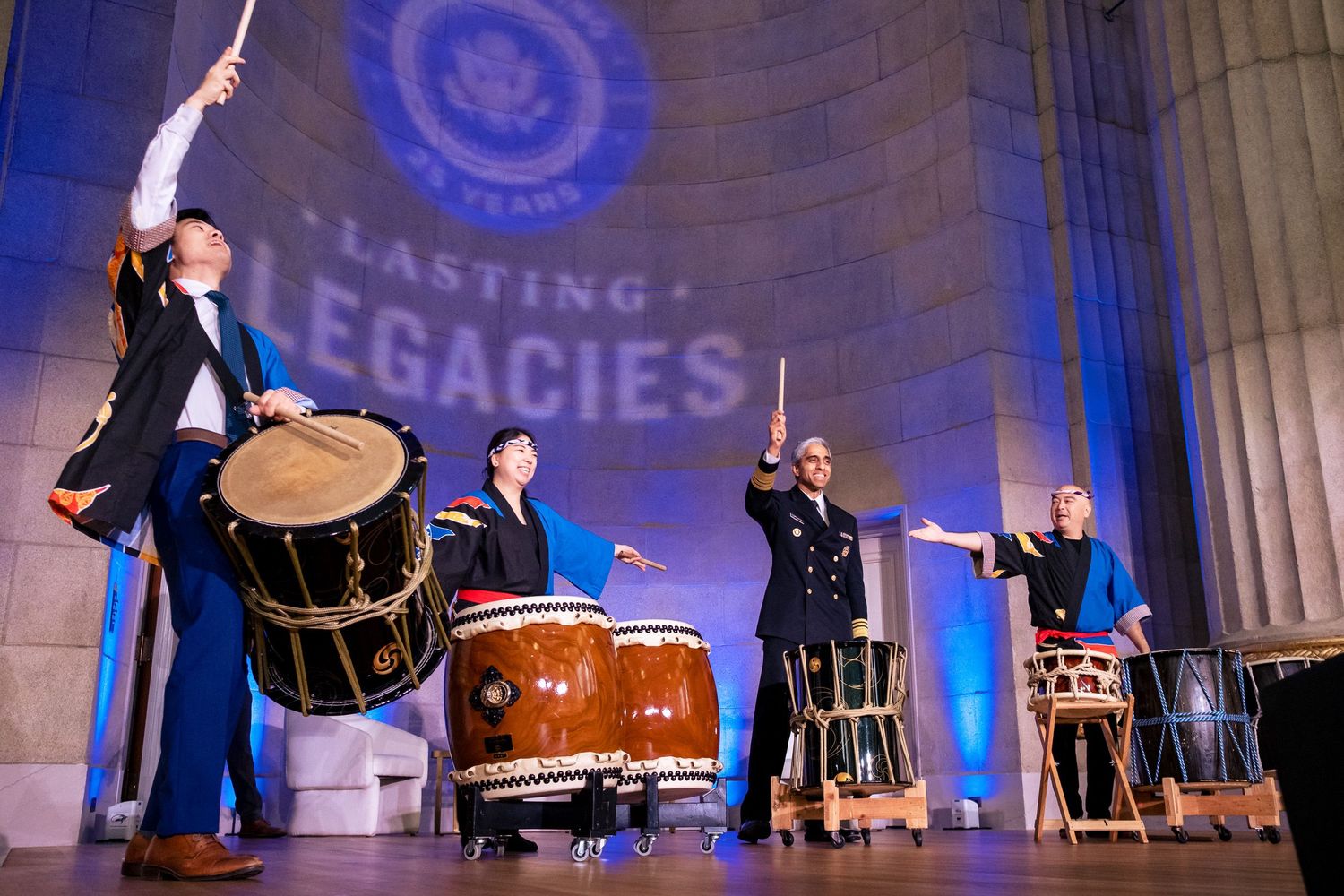 Taiko drummers perform on stage at a Lasting Legacies event, with blue lighting and a government seal backdrop.