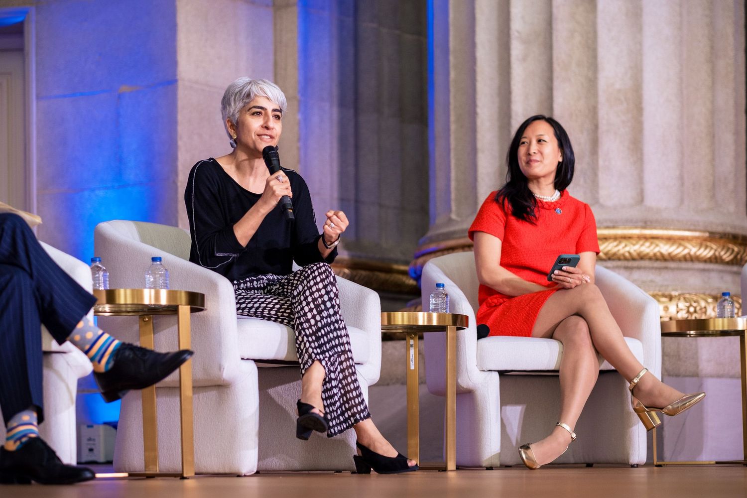 Two women speak on a panel at a formal event, one in black holding a microphone, another in red seated beside her.