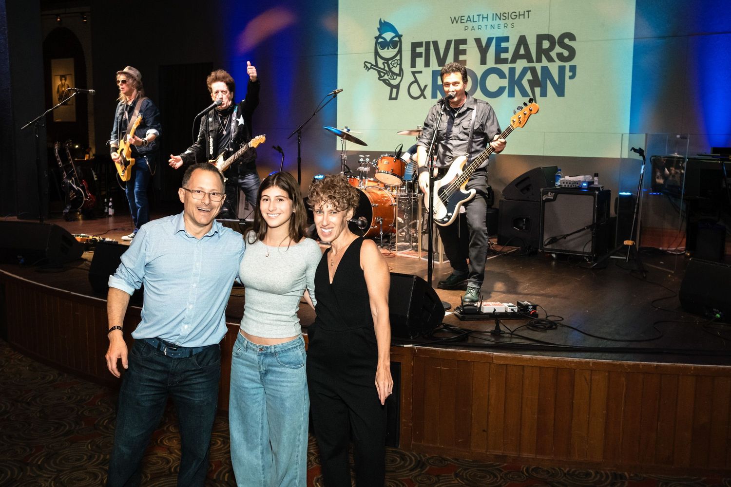 Three people pose smiling at Five Years & Kickin anniversary event with live band performing on stage.