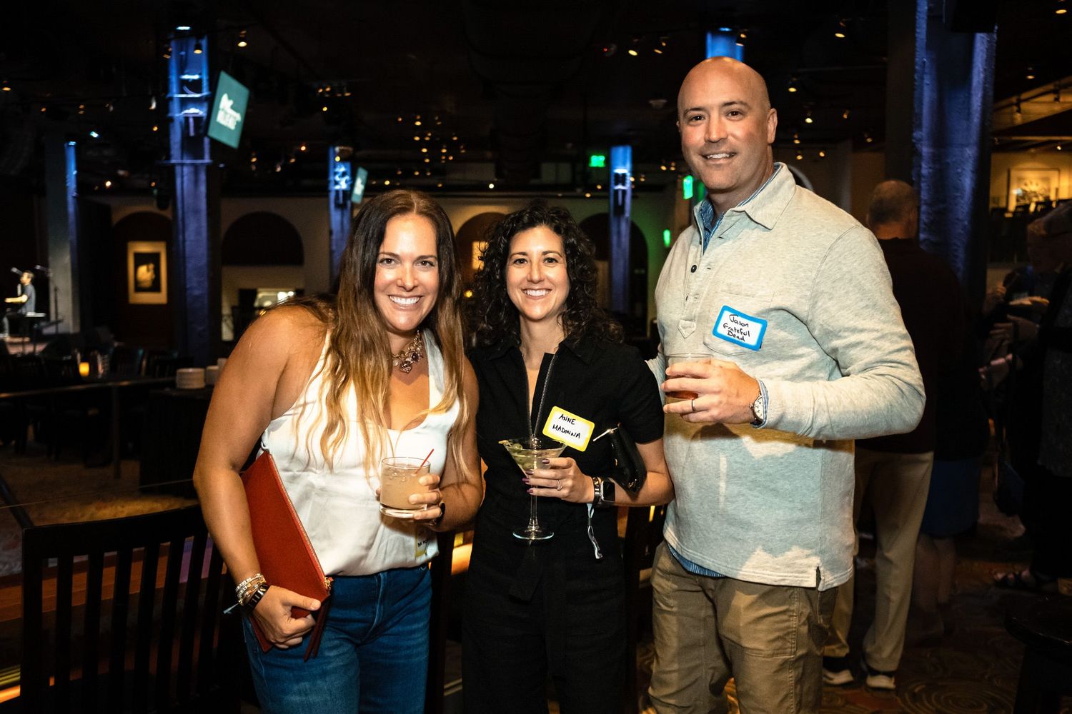 Three smiling professionals holding drinks at a networking event in a dimly lit venue.