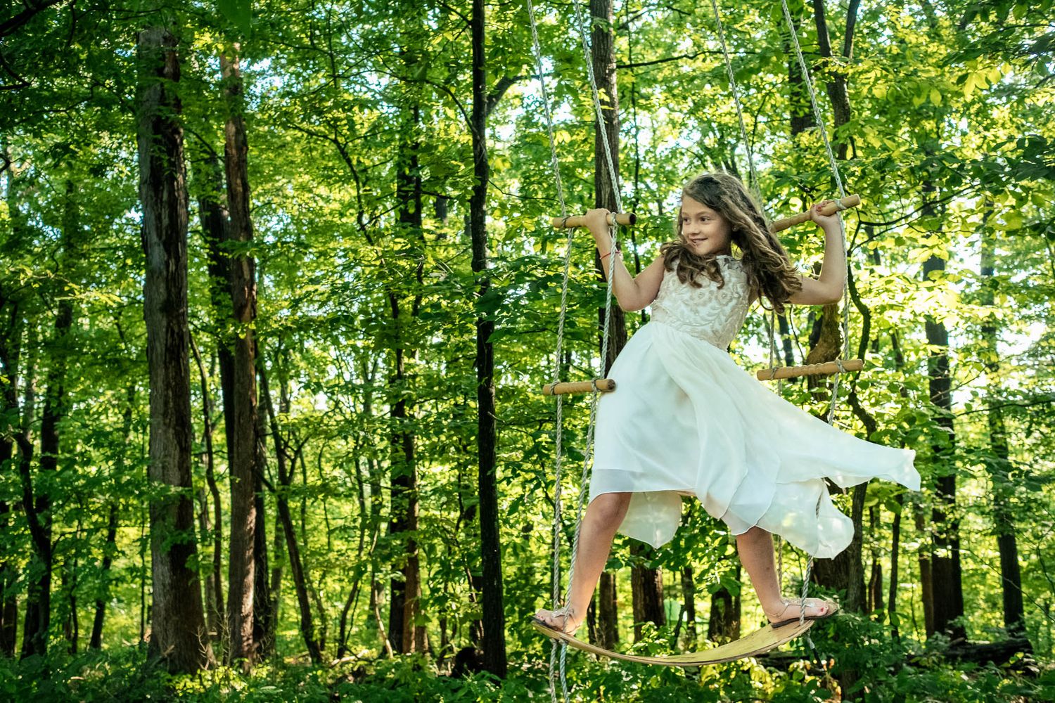 Young girl in white dress joyfully swinging on a wooden swing in a lush green forest.