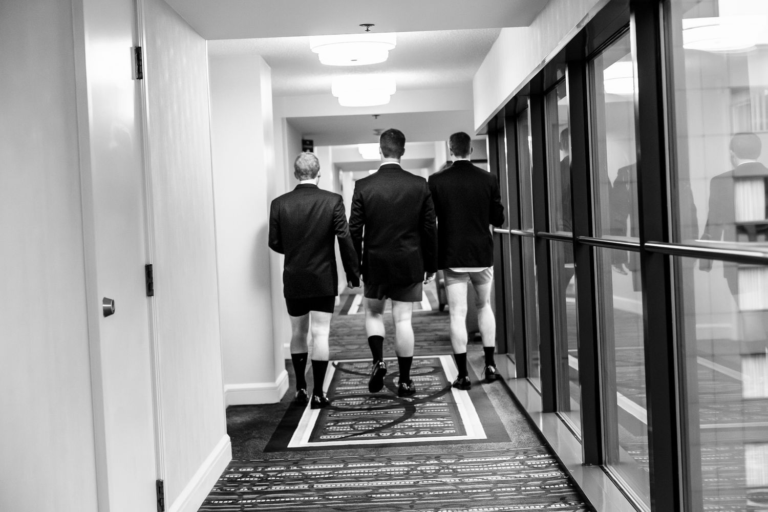 Black and white photo of groomsmen walking down a hotel hallway without pants before wedding.