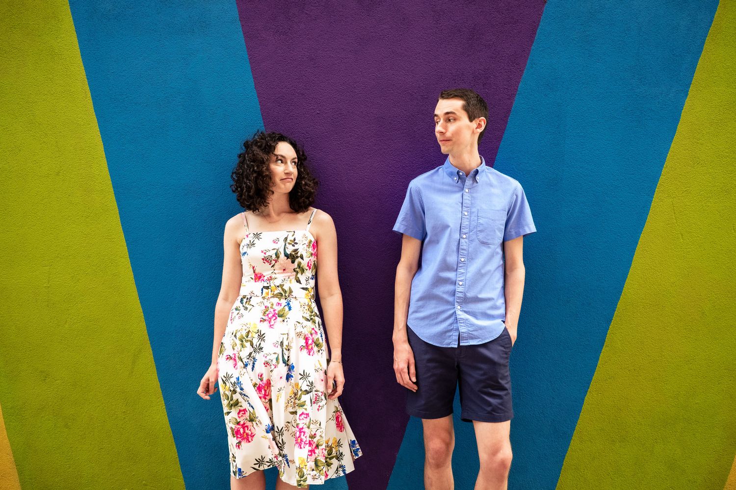 Couple stands apart smiling before colorful geometric mural wall.