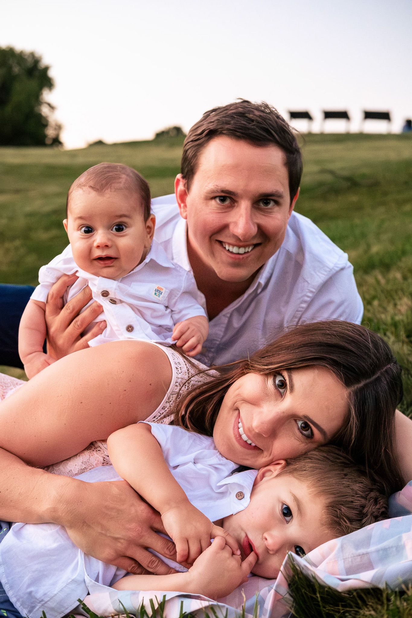 Happy young family of four lying in green grass field at golden hour sunset.