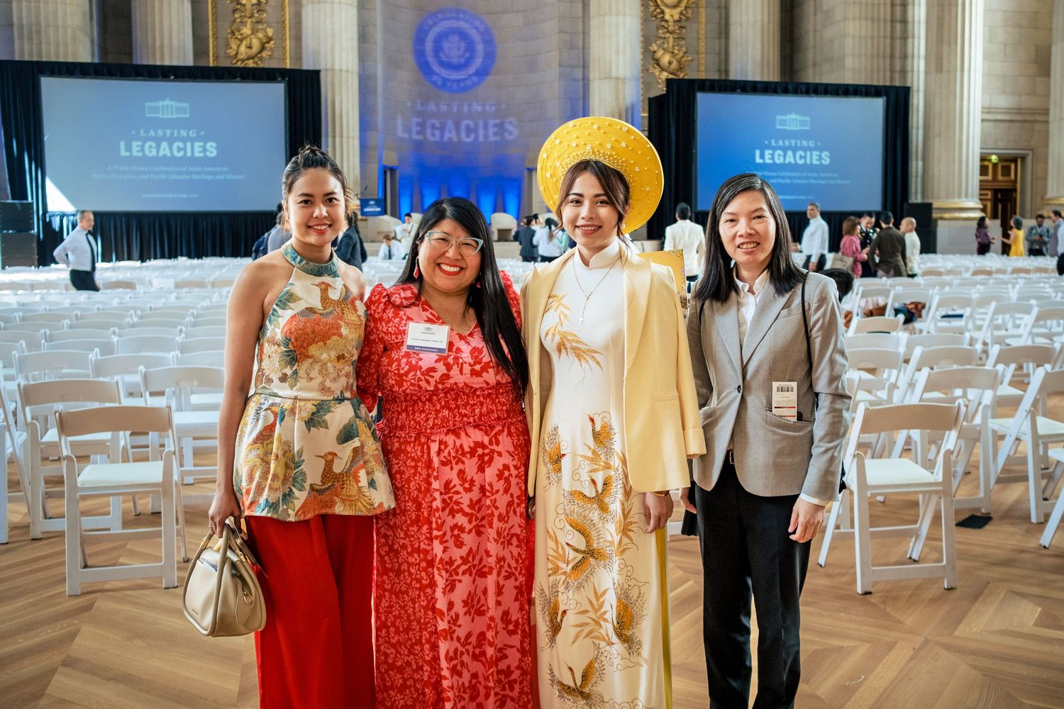 Four women in elegant attire pose together at a formal Legacies event in a grand hall.