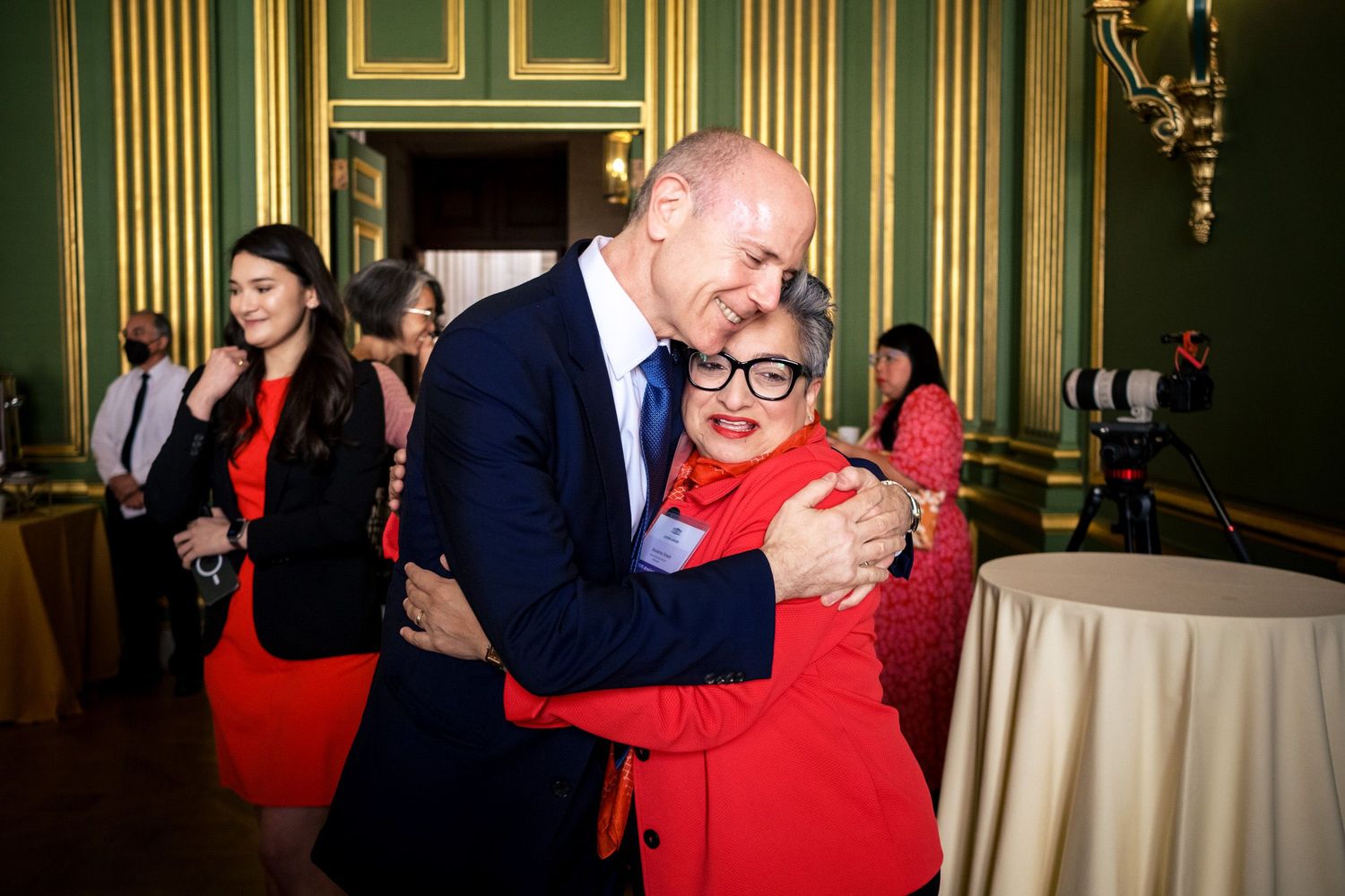 Two people share a warm hug in an elegant green-walled event hall, dressed in formal attire.