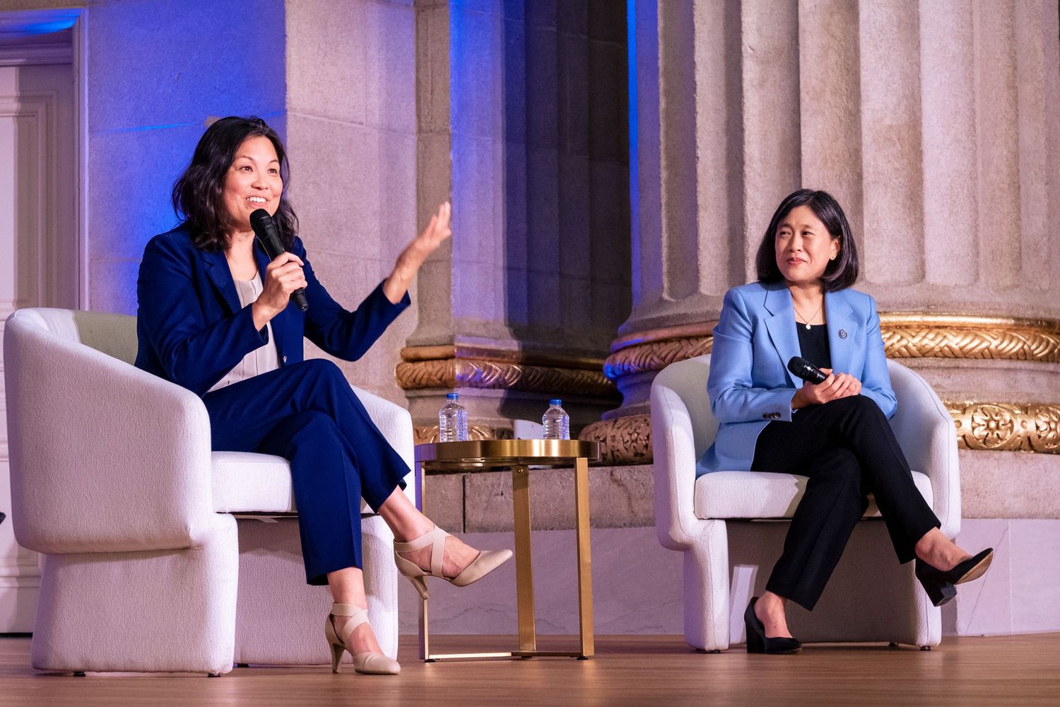 Two women in blue suits engage in a panel discussion on stage at a formal event, one speaking into a microphone.