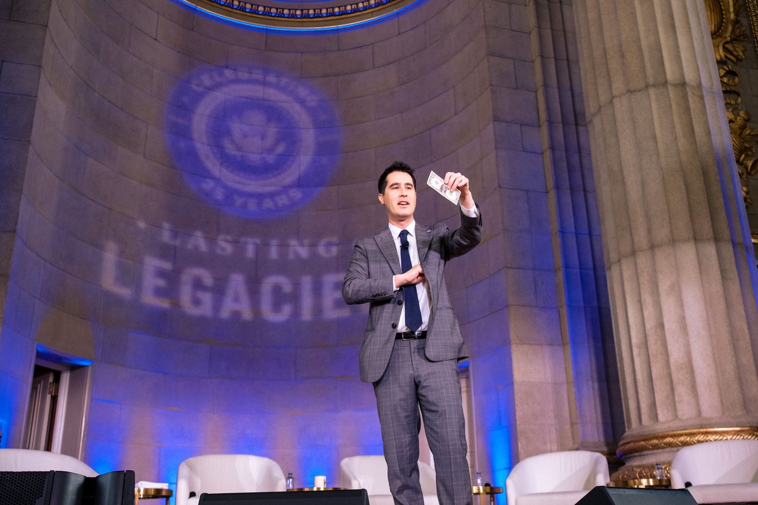 Man in gray suit holds paper on stage at Lasting Legacies event with blue lit columns.