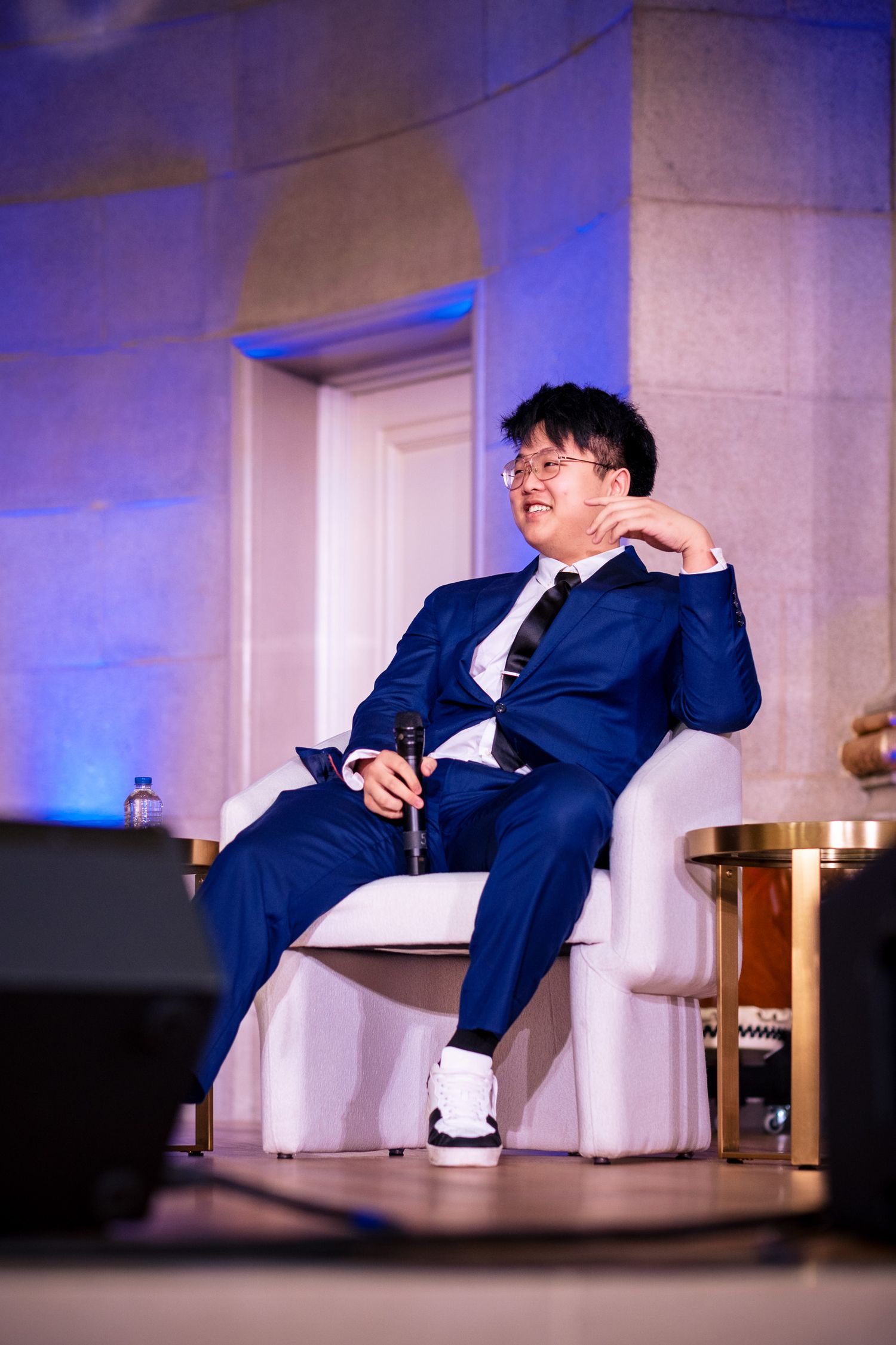 Man in blue suit seated relaxed in white chair on stage at formal conference event.