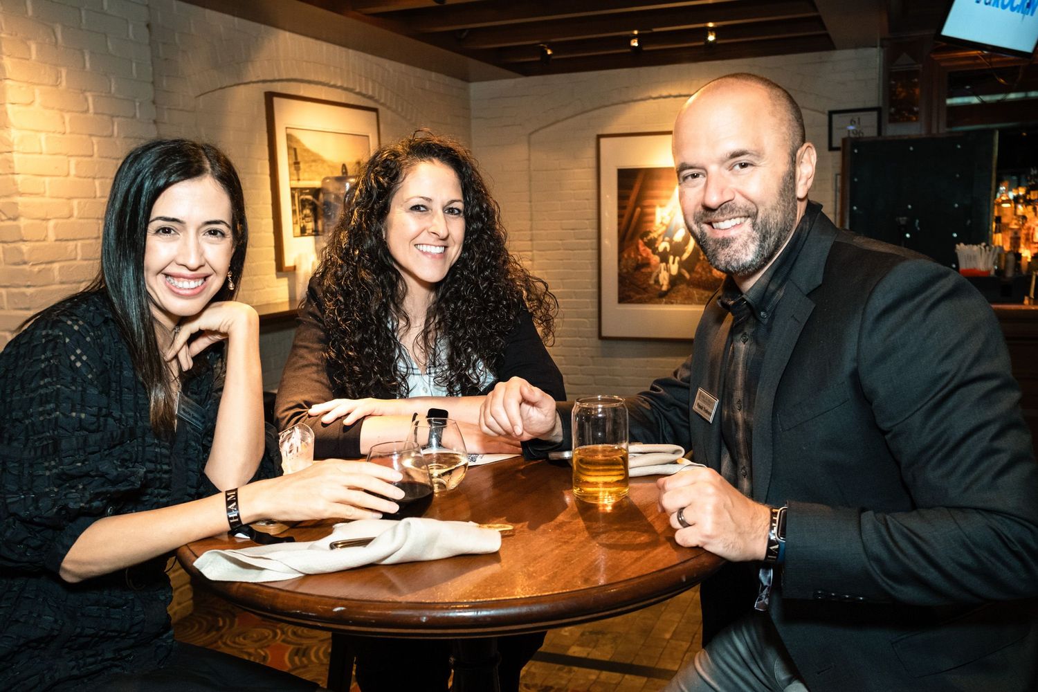 Three friends smiling at a round wooden table in a cozy restaurant, enjoying drinks together.