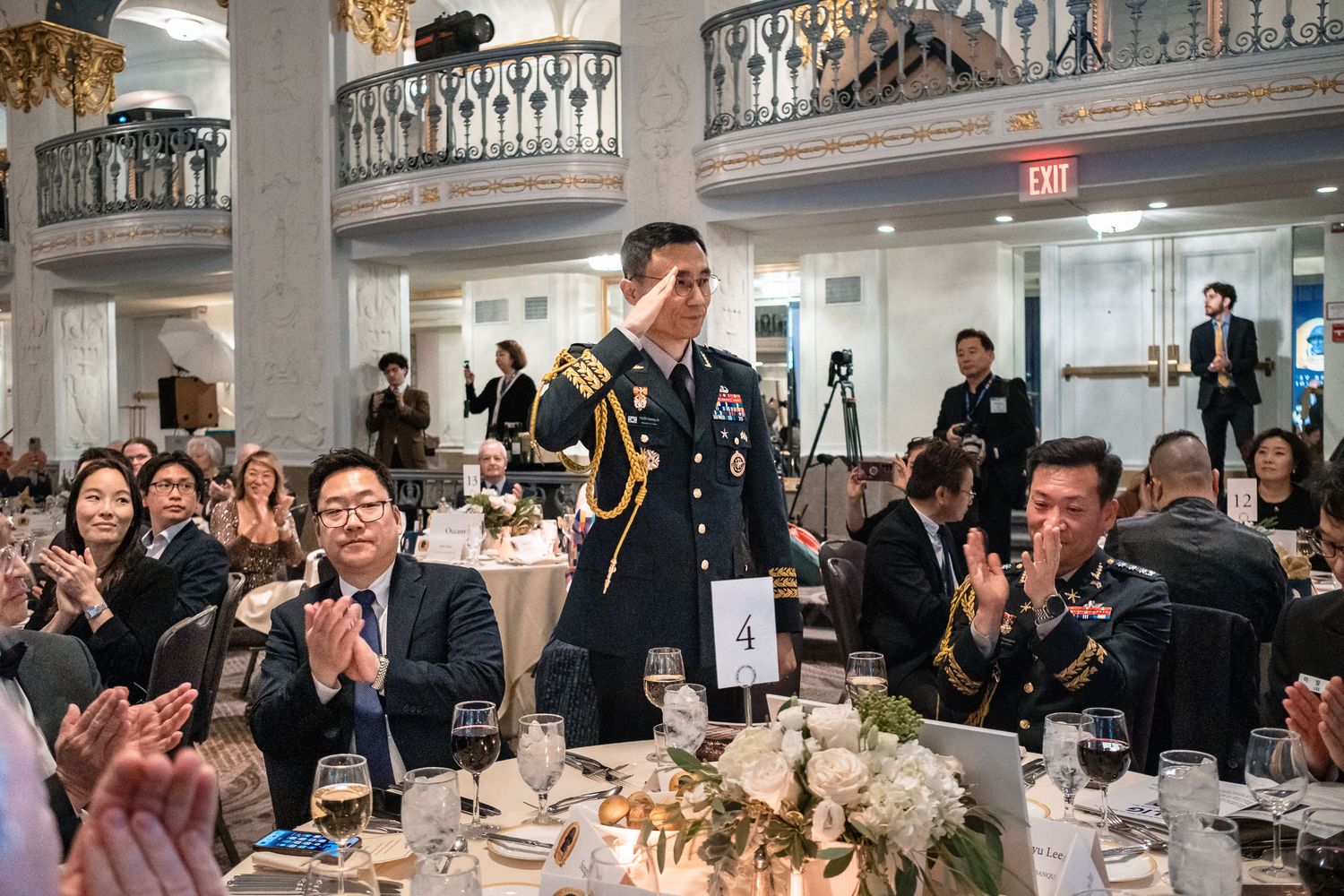 Military officer in dress uniform salutes at formal gala dinner, surrounded by applauding guests in elegant ballroom.