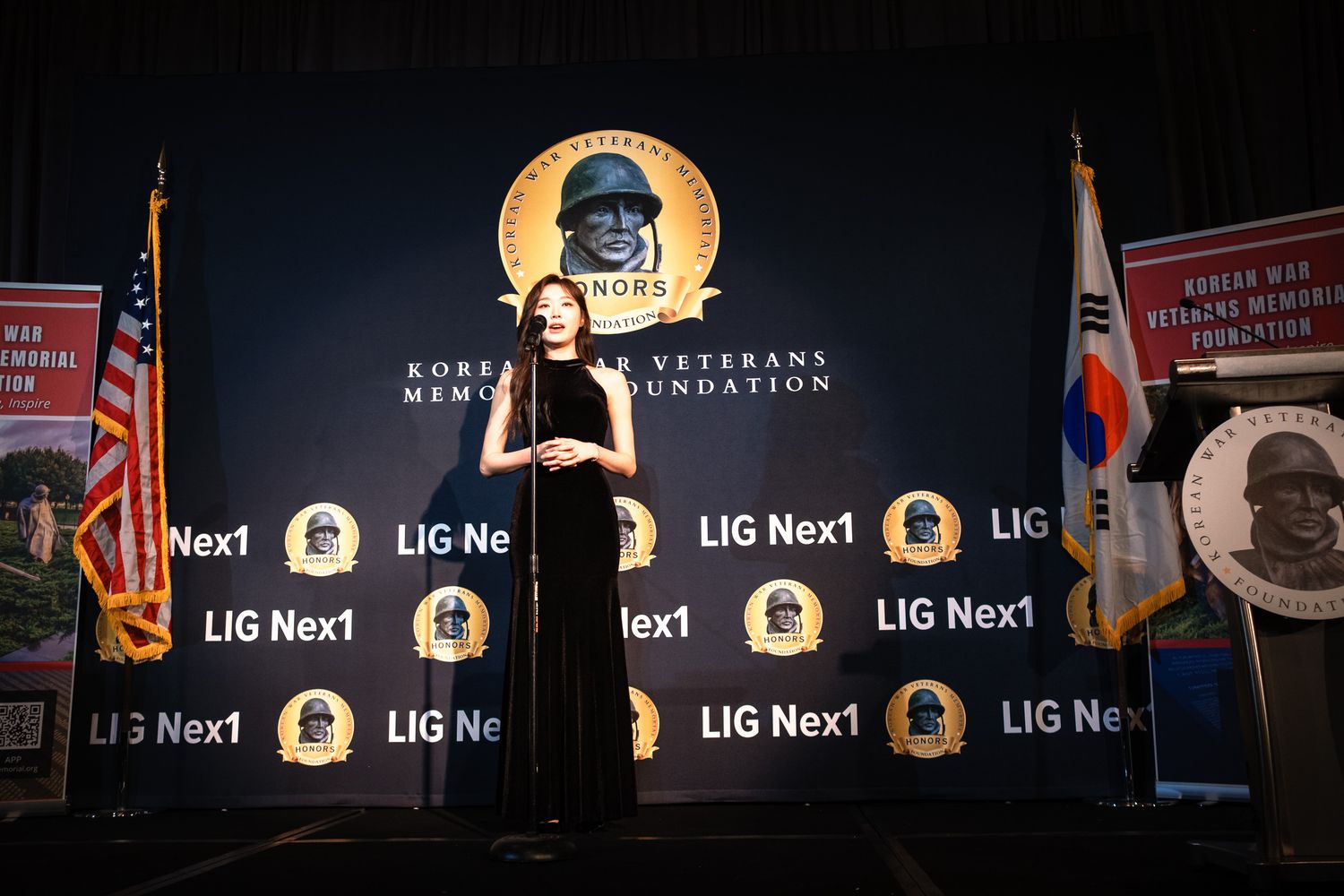 Woman speaks at Korean War Veterans Memorial Foundation event with LIG Nex1 branded backdrop and flags on stage.