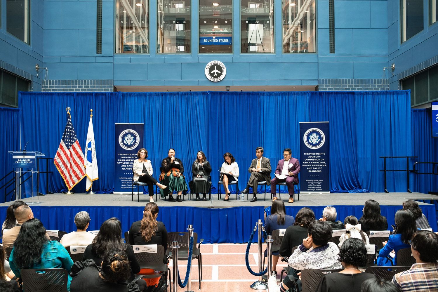 Panel of speakers on a blue-draped stage address an audience at a formal government conference event.