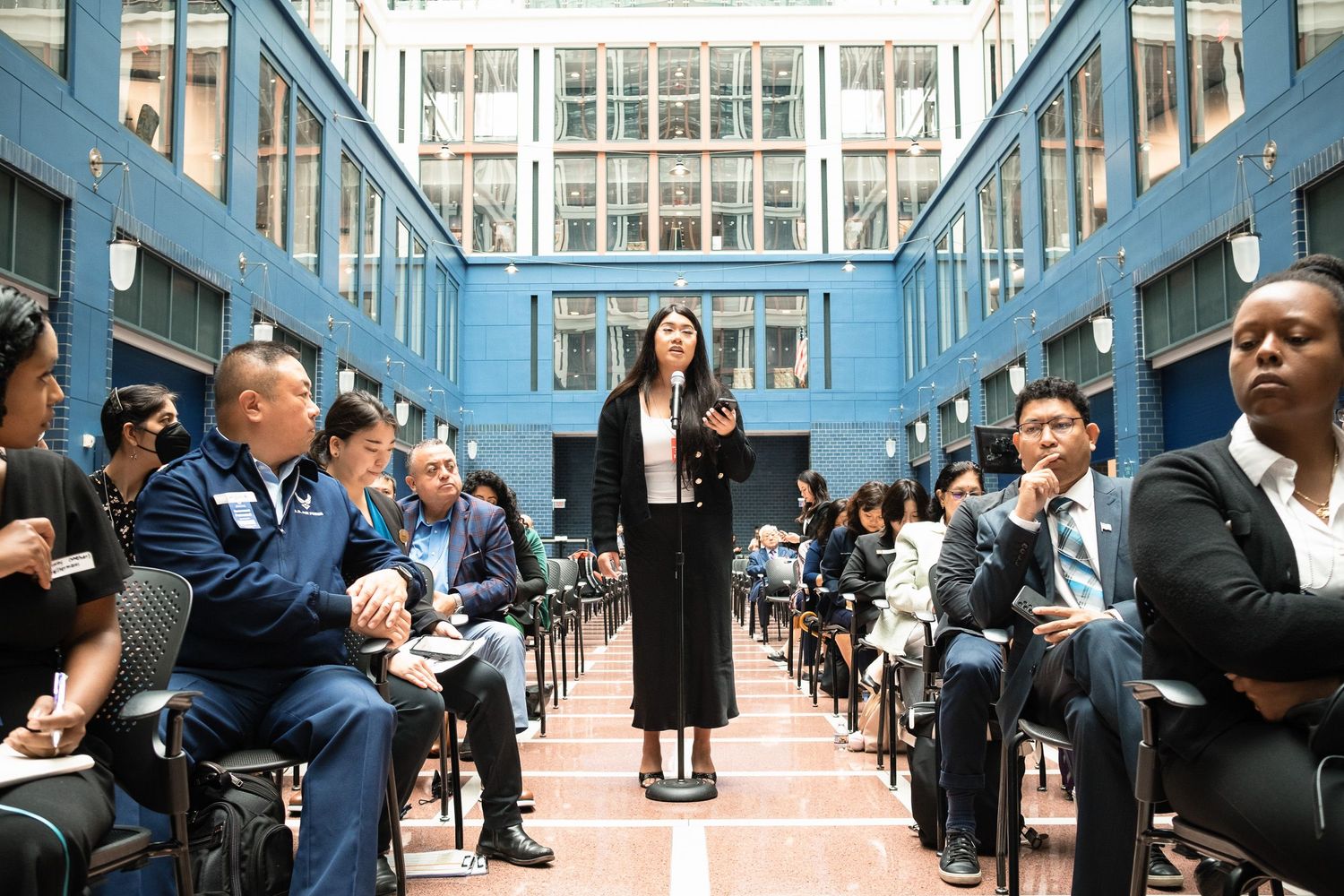 Woman speaking at a formal indoor event with seated audience in a blue-walled atrium venue.