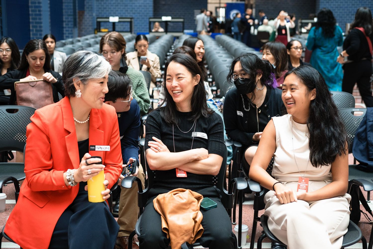 Three women laugh together at a professional networking event in an auditorium filled with diverse attendees.