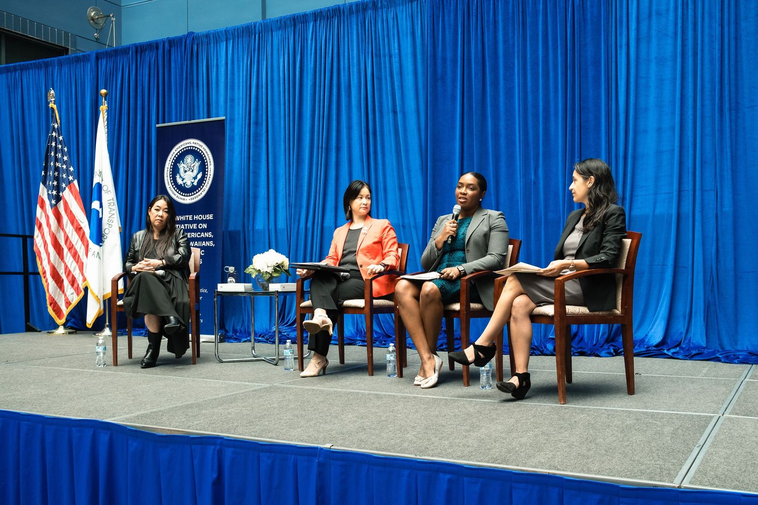 Four women participate in a panel discussion on stage with US and government flags at a formal conference event.