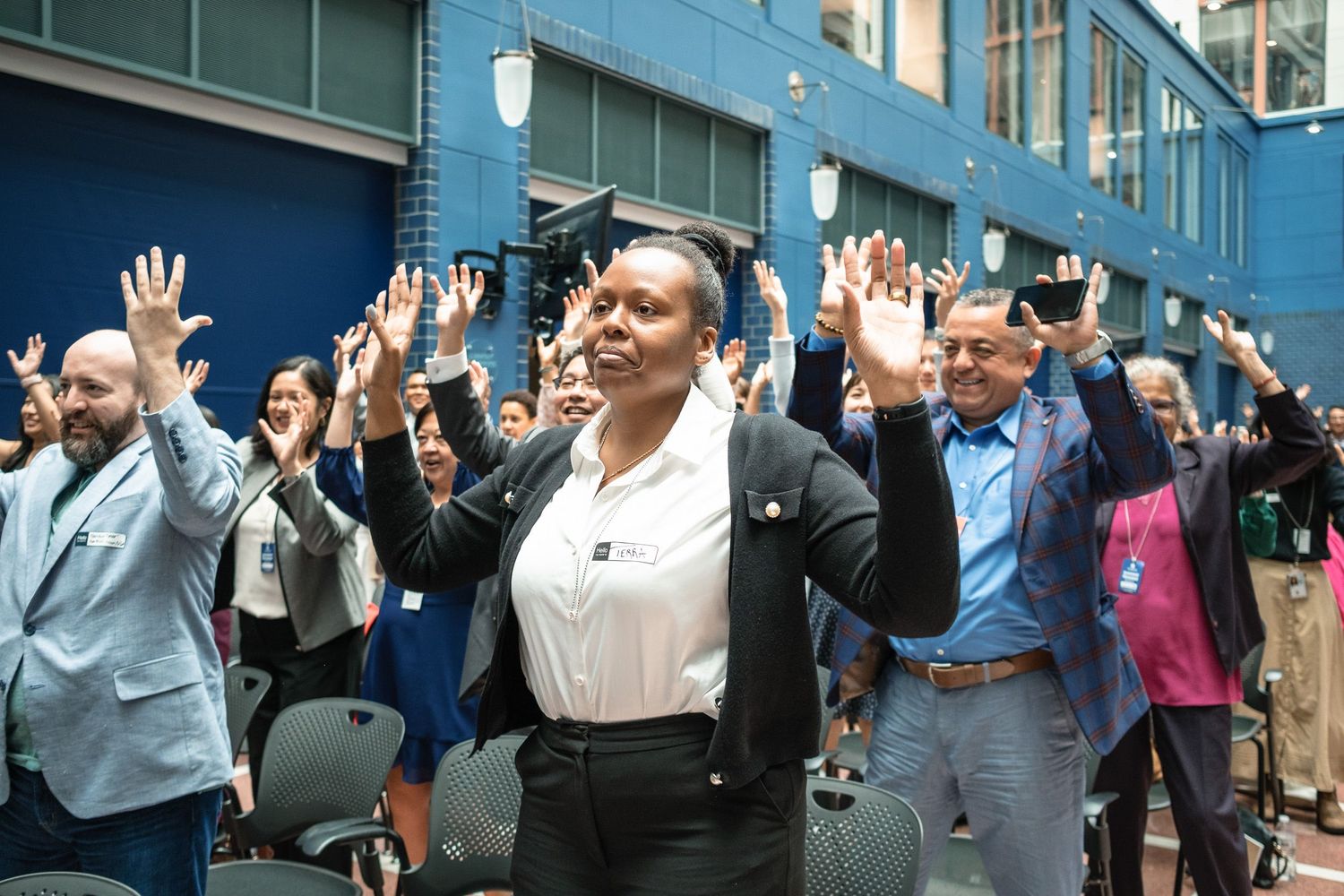 Diverse group of people raising hands enthusiastically at an indoor event or conference gathering.