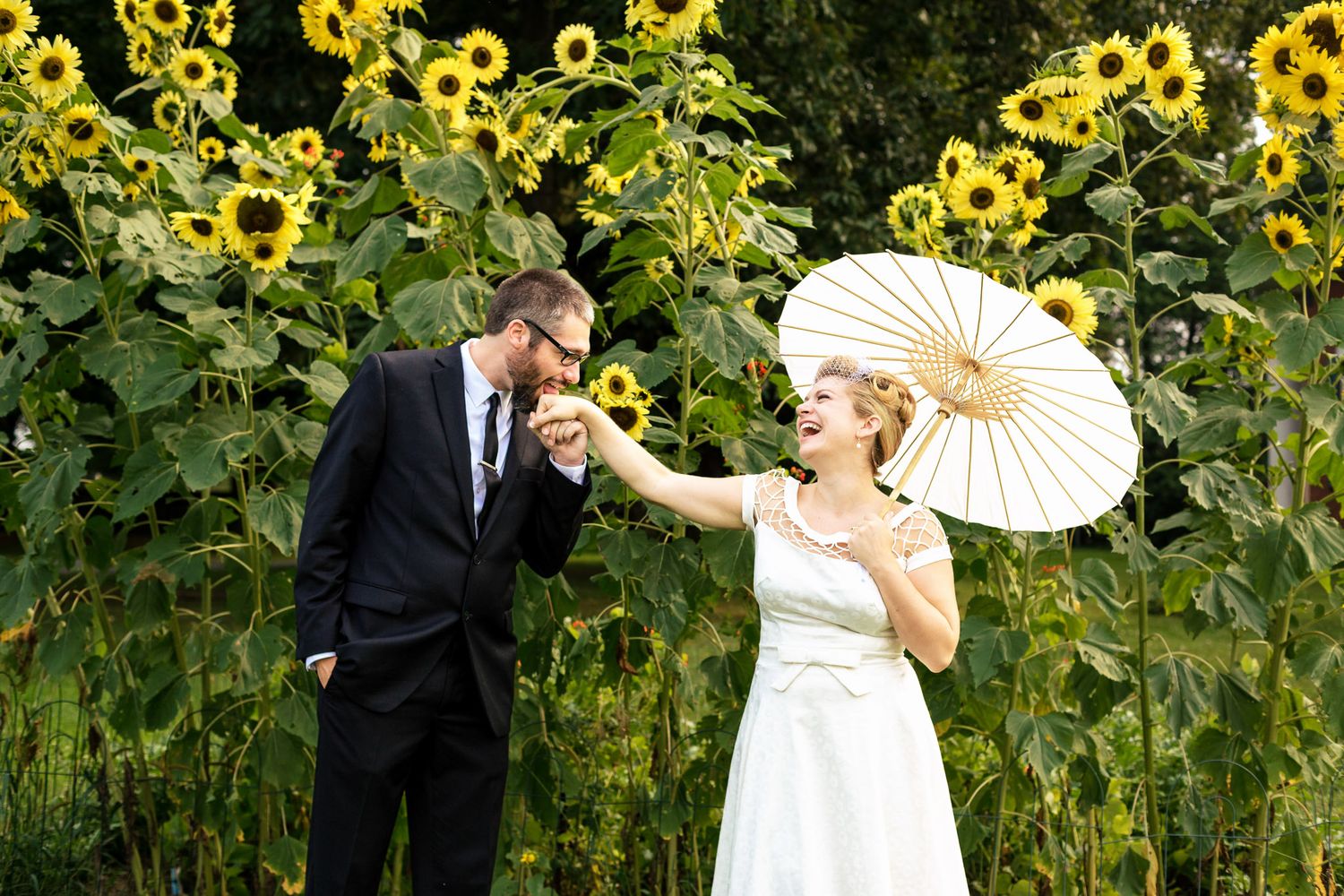 Bride holding parasol while groom kisses her hand in a sunflower field wedding photo.