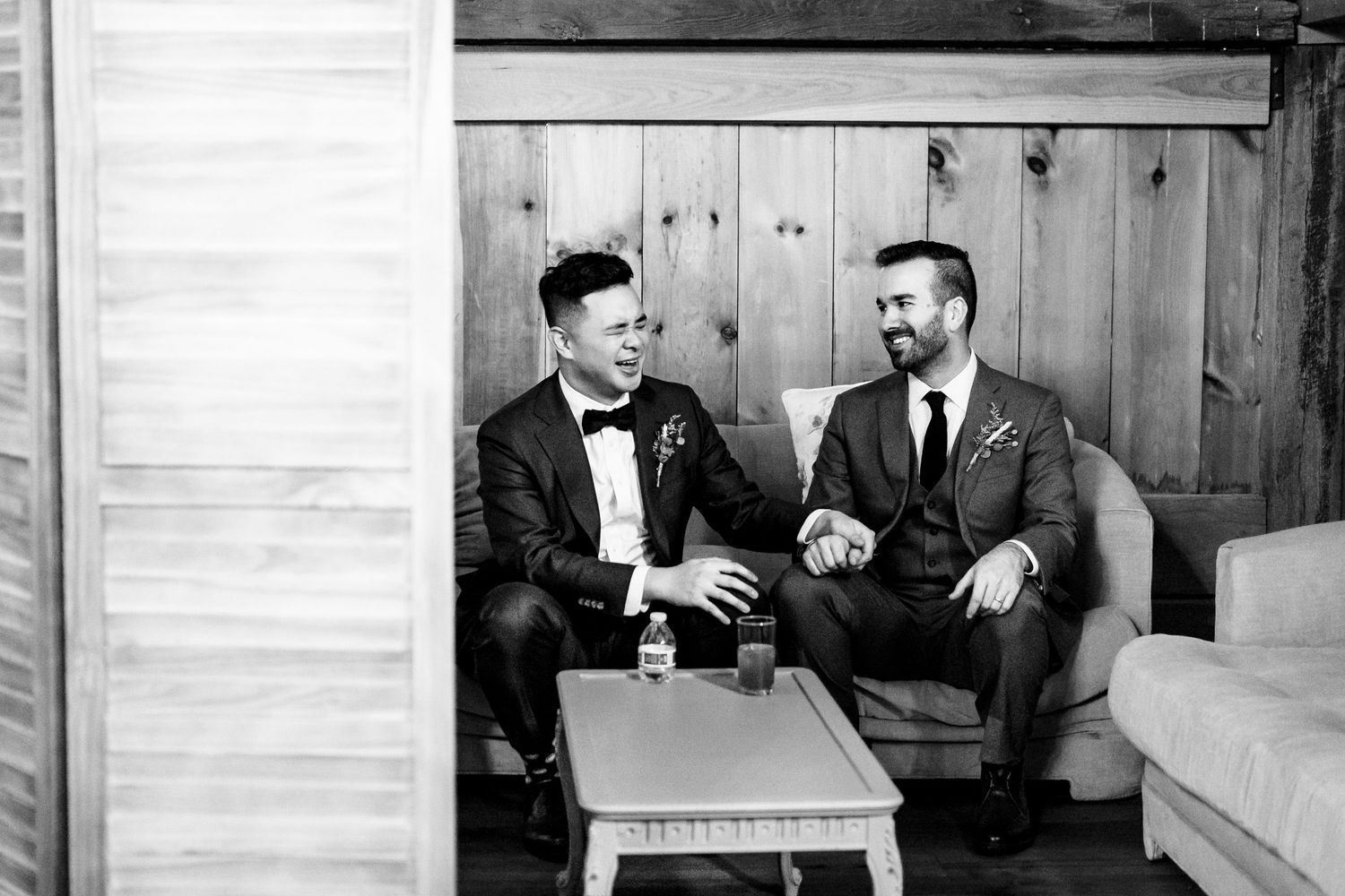 Black and white photo of two grooms sharing a private moment on a couch before wedding ceremony.