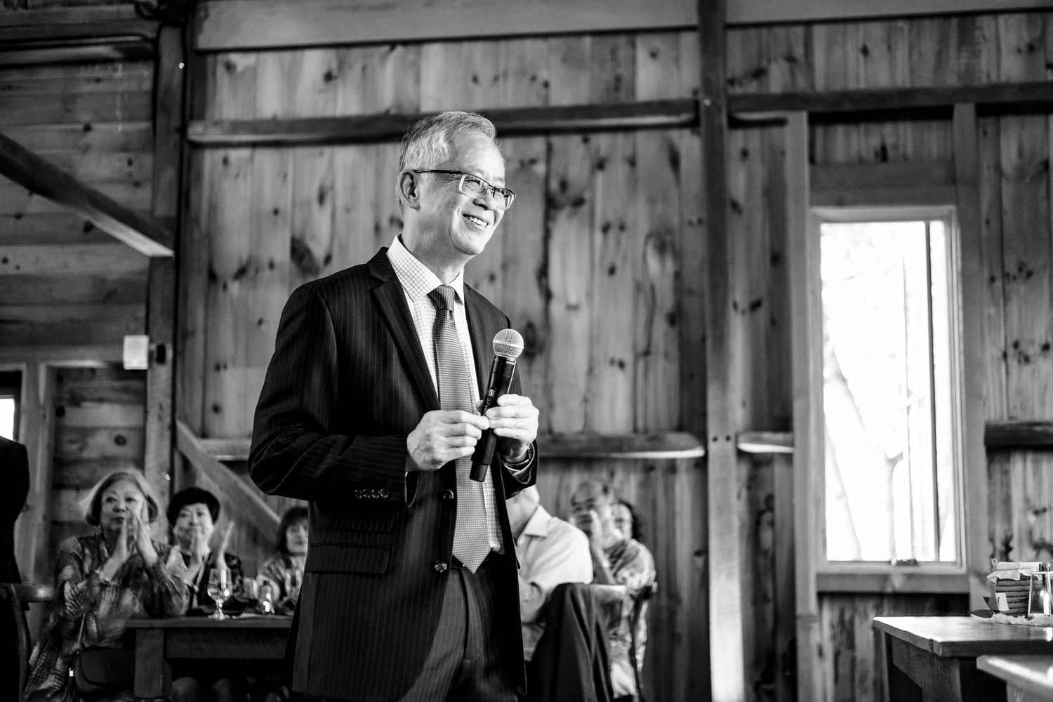 Man in dark suit giving a speech at a rustic barn wedding reception while holding a microphone.
