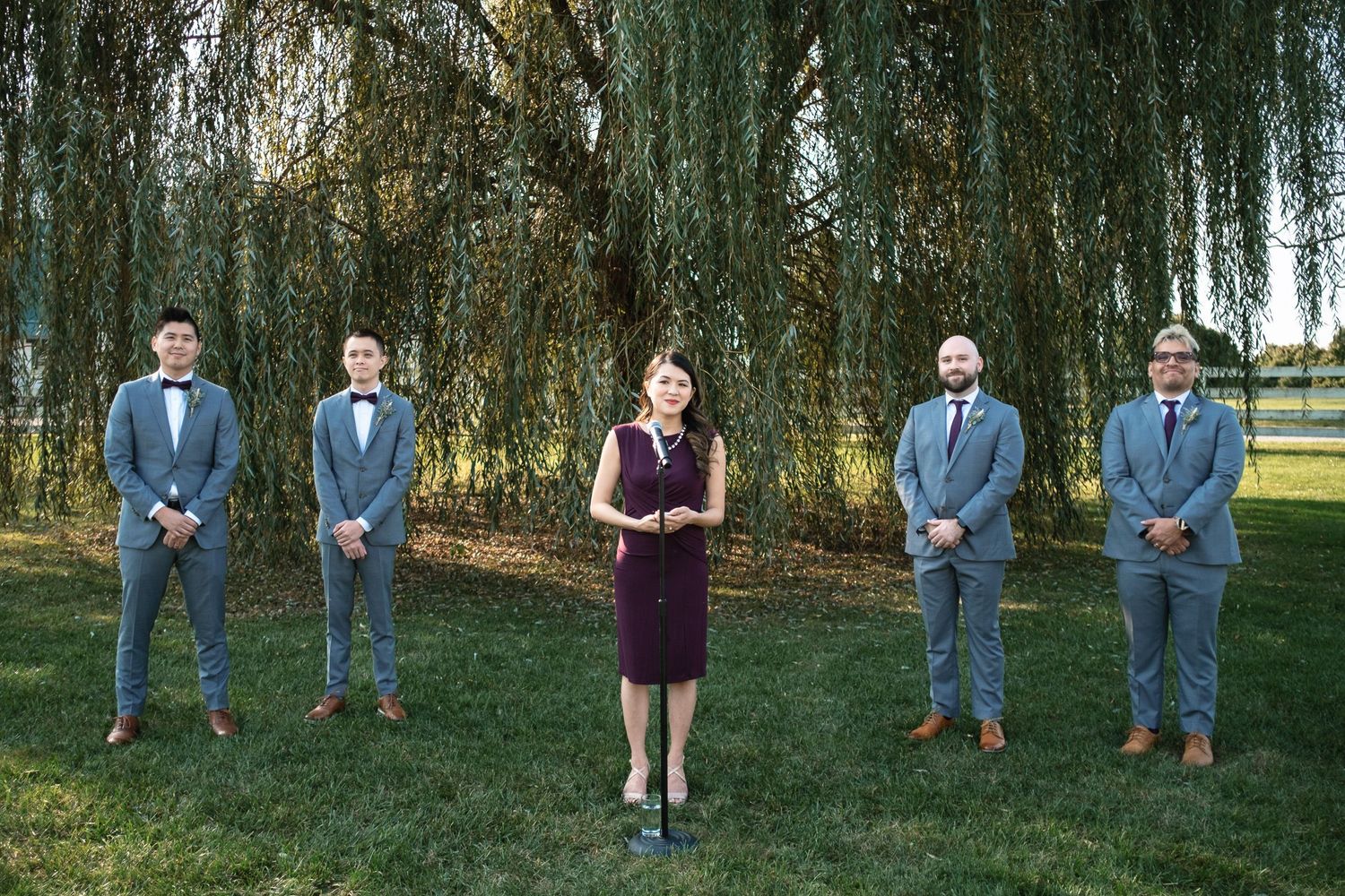 Wedding officiant in burgundy dress stands with four groomsmen in gray suits under a weeping willow tree.