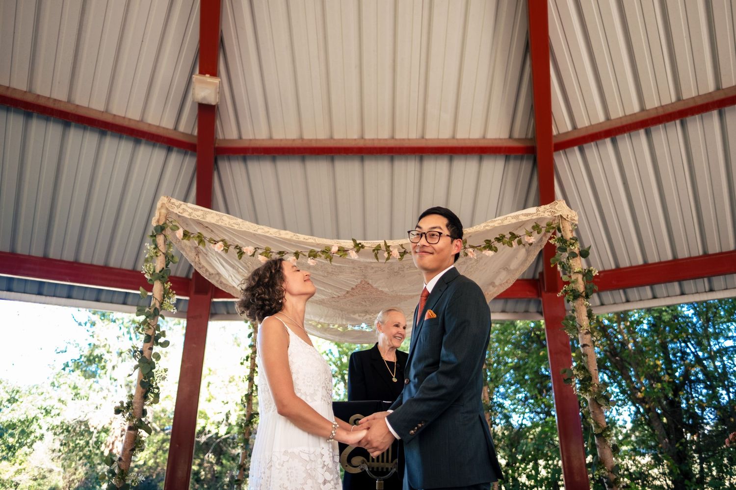 Bride and groom hold hands at altar under floral draped arch with officiant during wedding ceremony.