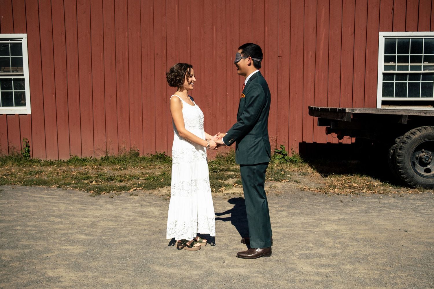 Bride in white dress and groom in dark suit holding hands near red barn.