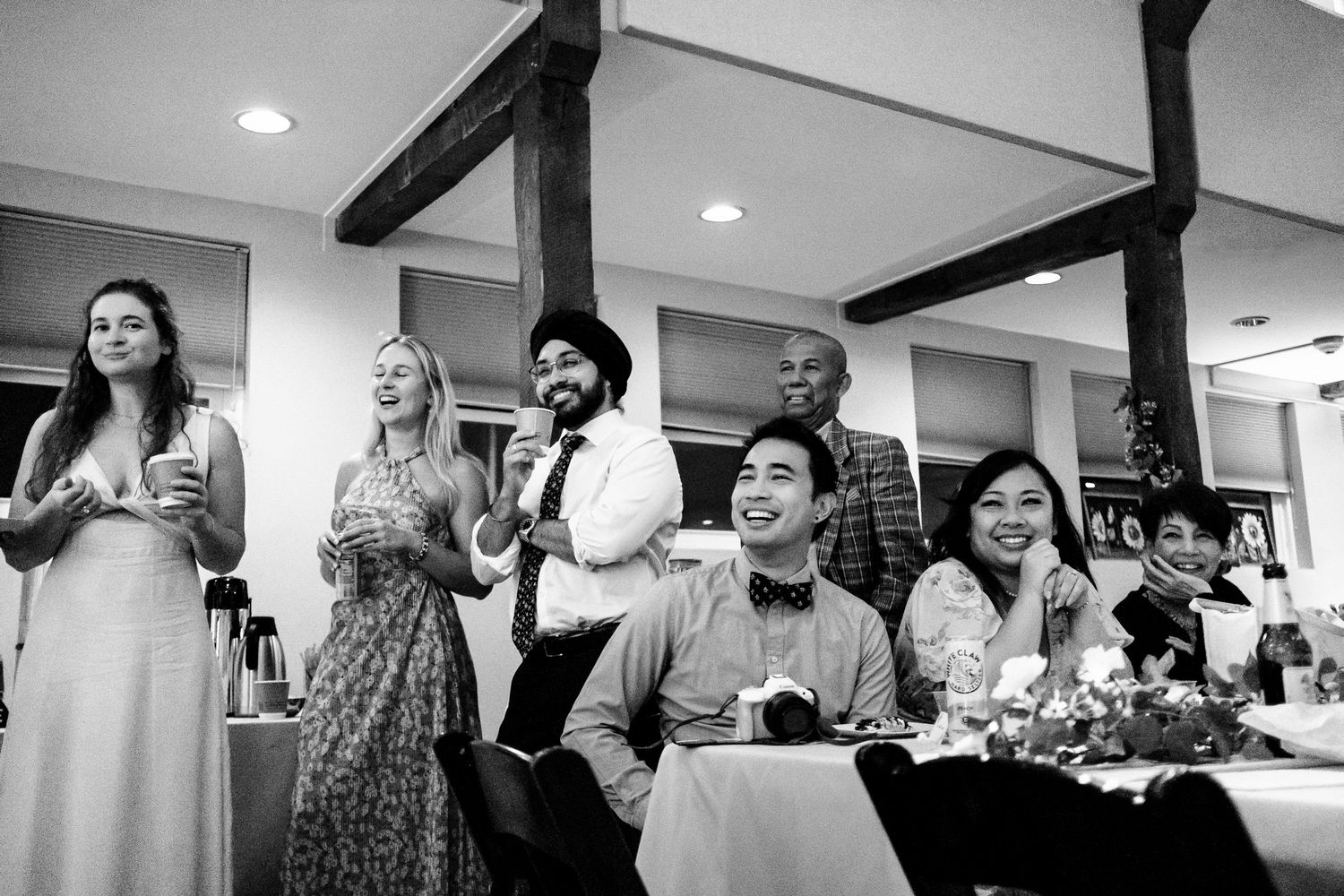 Diverse wedding guests laugh and celebrate during a speech at a reception dinner in black and white.