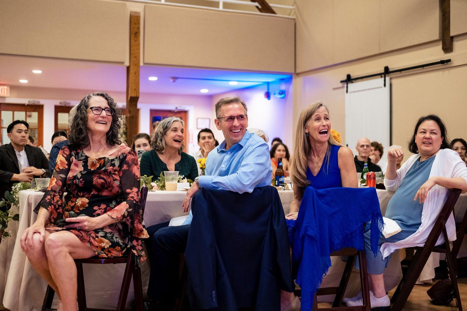 Wedding guests laugh and smile joyfully while seated during a lively wedding reception celebration.