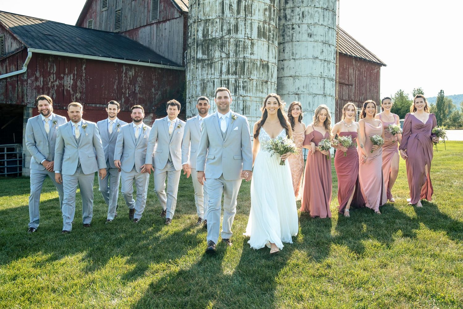 Bride and groom lead the full wedding party walking near a rustic red barn and silo.