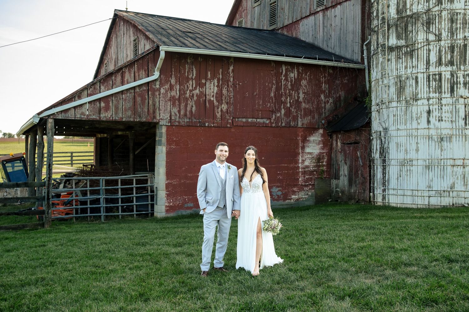 Bride and groom stand together on green lawn beside a rustic red barn and stone silo.