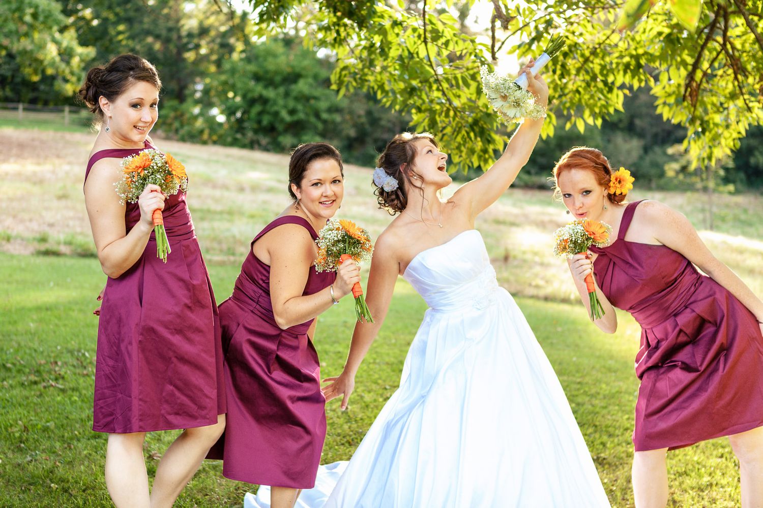 Bride and three bridesmaids in burgundy dresses laugh and pose outdoors in a park.