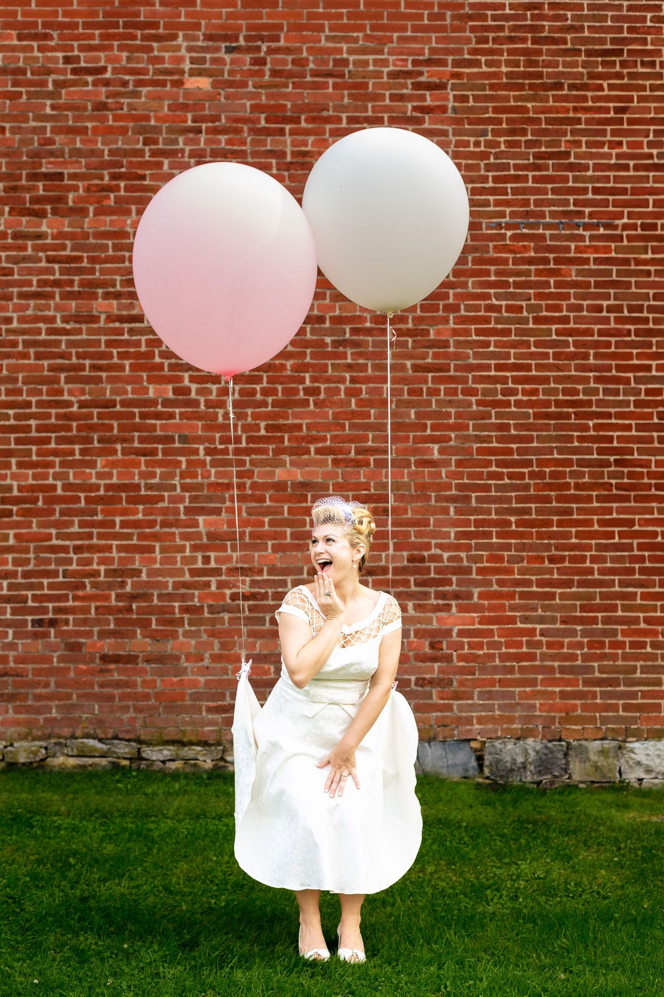 Bride in white dress holding pink and white balloons in front of a red brick wall.