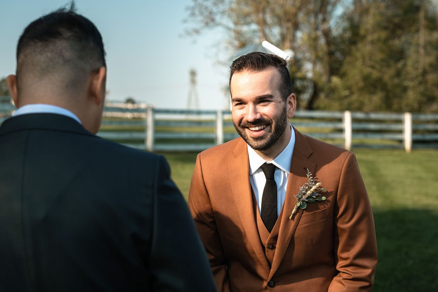 Groom in rust brown three-piece suit smiling warmly at his partner during outdoor wedding ceremony.