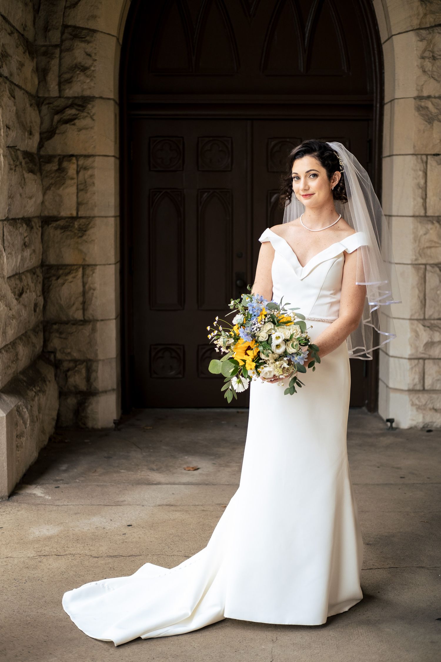Elegant bride in off-shoulder white gown with veil holds colorful bouquet before stone church doors.
