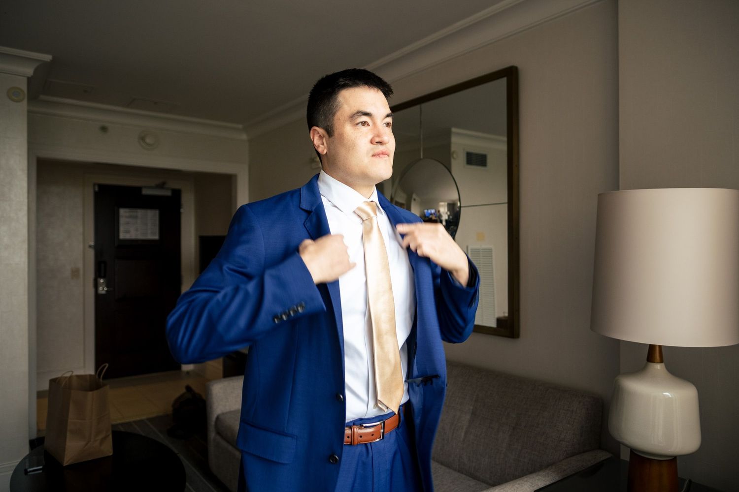 Groom in royal blue suit adjusting his tie in a hotel room mirror before wedding.