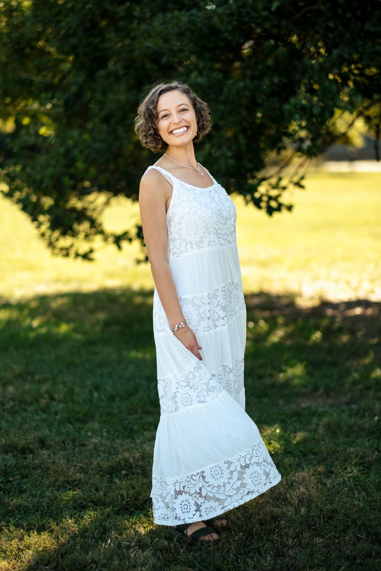 Smiling bride in a white lace maxi dress standing in a sunlit garden with lush green trees behind her.