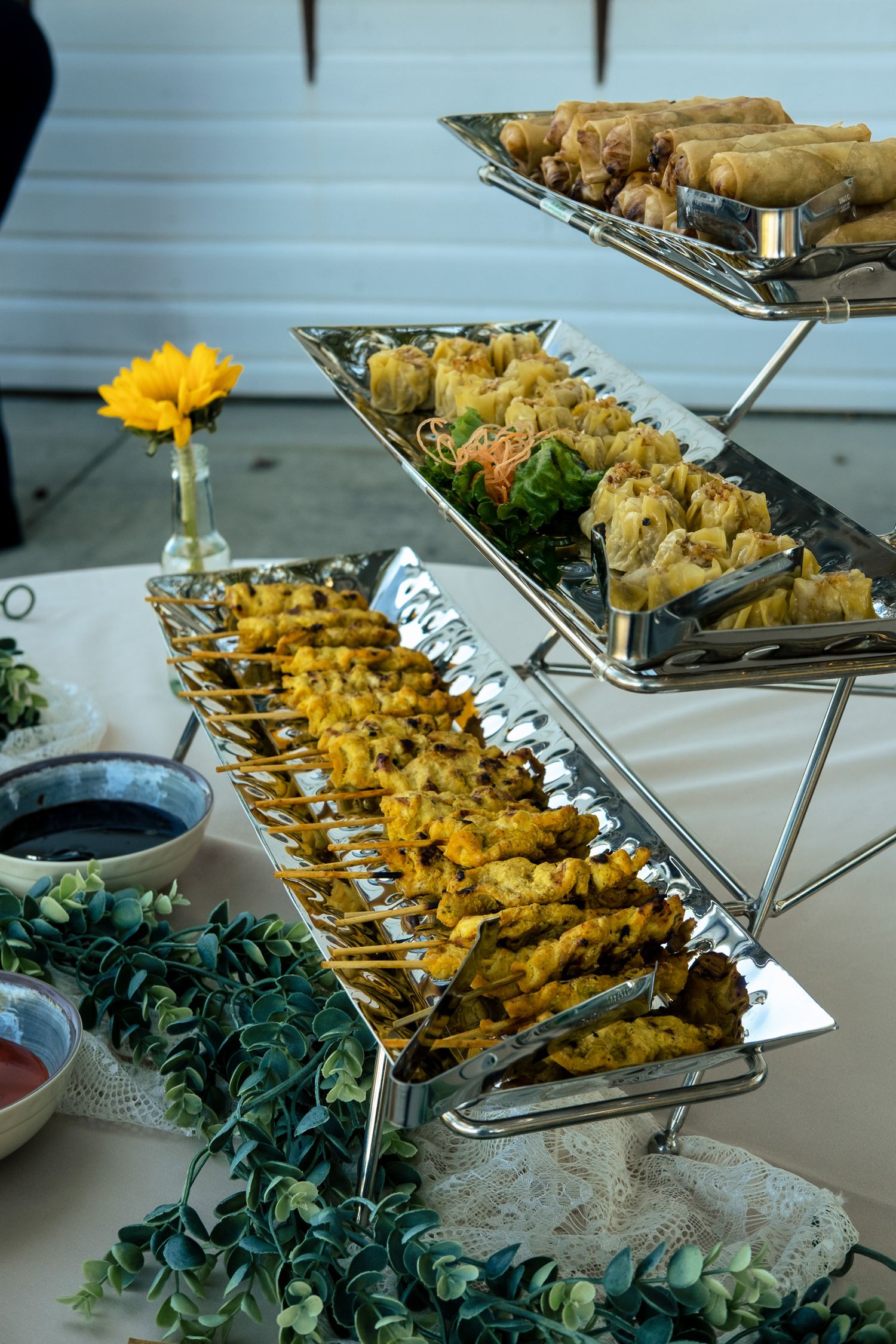 Elegant three-tier wedding appetizer display with skewers, spring rolls, and dipping sauces.