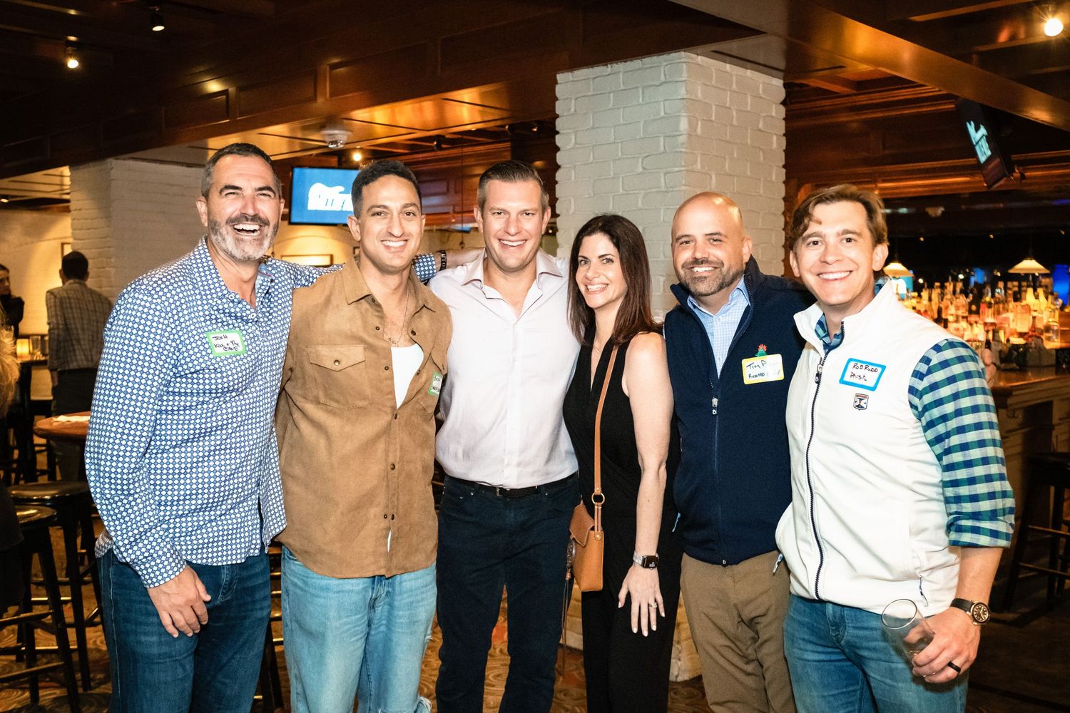 Six people smiling and posing together at a casual indoor networking event or party near a bar.