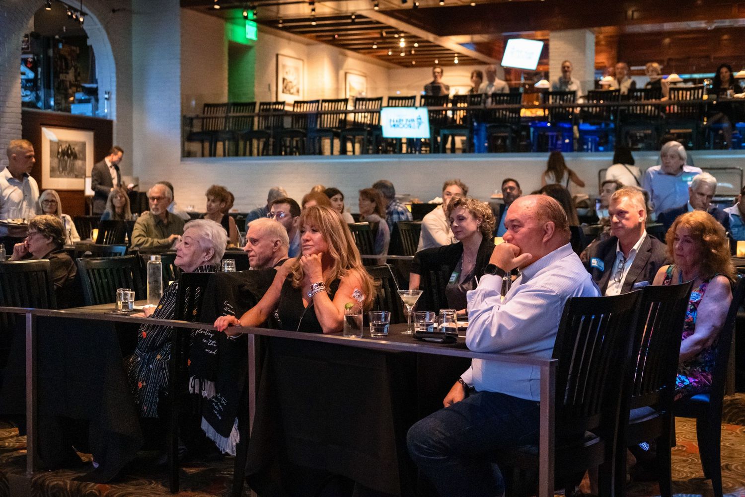 Audience members seated at tables in an upscale restaurant venue attentively watching a presentation or event.