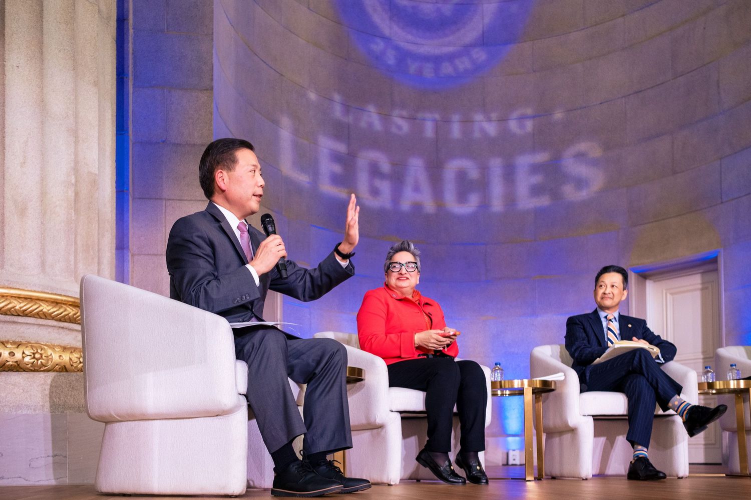 Three panelists discuss on stage at a Lasting Legacies event beneath an ornate backdrop.