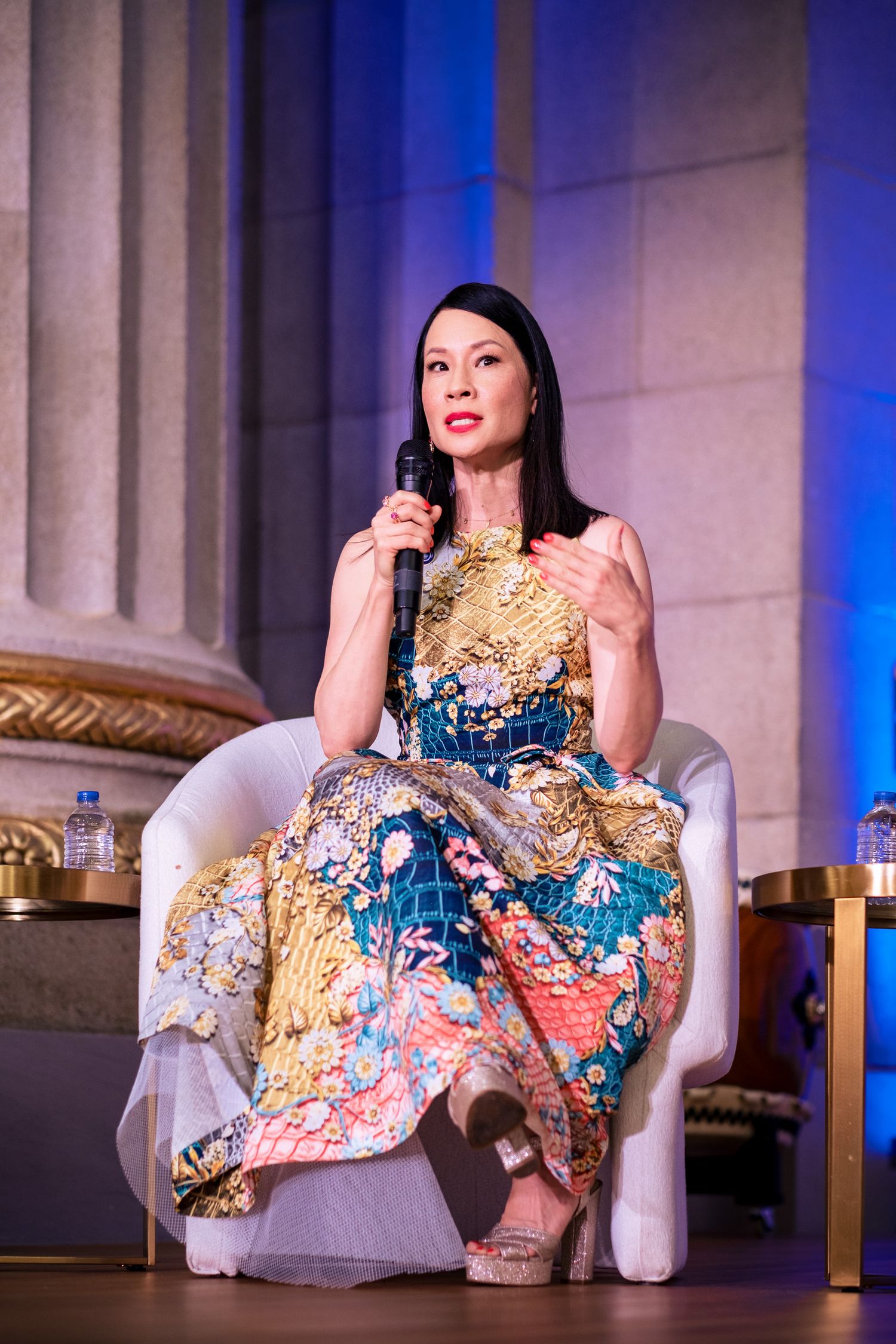 Woman in colorful floral gown speaks into microphone while seated on stage at formal event.