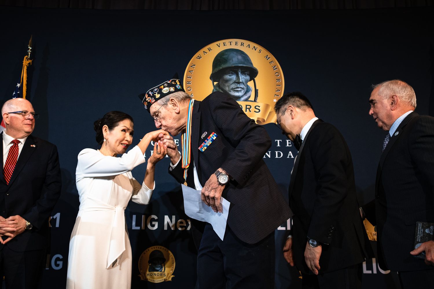 Woman in white dress congratulates award recipient on stage at formal military veterans ceremony.